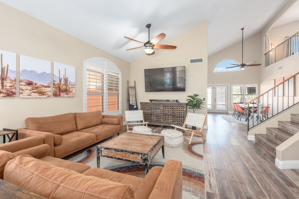 A spacious living room with a large brown leather sofa, a coffee table, and a flat-screen TV mounted on the wall. There are ceiling fans, a staircase, and a dining area with orange chairs.