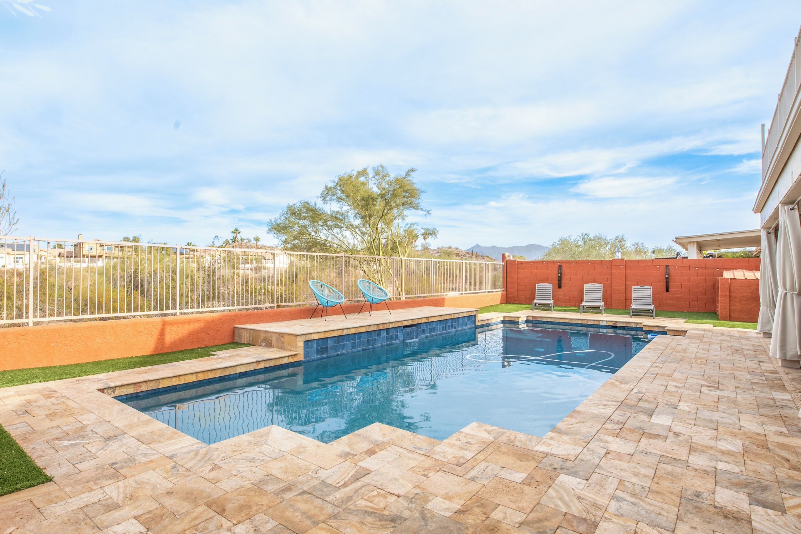 A swimming pool with a tiled deck, two blue chairs, and a fenced area with a red building in the background.