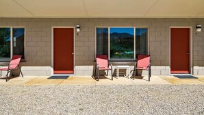 Un motel avec deux chambres, chacune dotée d’une porte rouge et d’une fenêtre. Devant chaque chambre, on trouve deux chaises et une petite table.