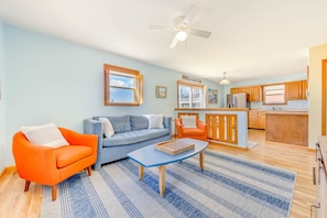 A living room with a sofa, armchairs, a coffee table, and a kitchen area with wooden cabinets and a refrigerator. - Soundside Dog Friendly Home w/ Hot Tub, Smart TVs, and Nearby Beach Access (Avon)