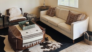 Un salon chaleureux avec un plafond en bois, un canapé blanc, un fauteuil en cuir marron, une petite table d’appoint avec une lampe et un tapis en peau de vache.