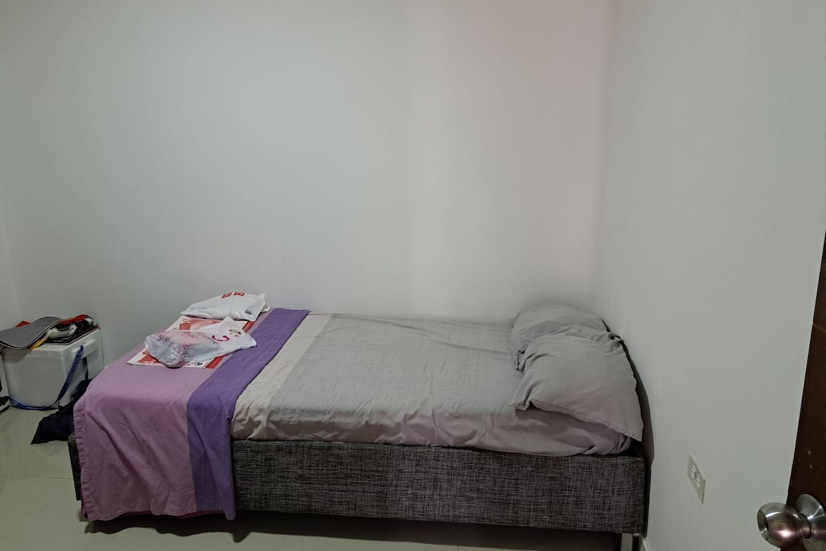 A small, single-bed room with a grey mattress and a purple blanket. There is a small table with a chair to the left, and a door with a silver handle on the right.