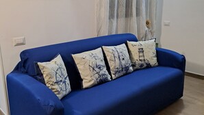 Un canapé bleu agrémenté de coussins sur le thème de la marine, un tableau représentant un paysage côtier accroché au mur et un rideau à rayures.