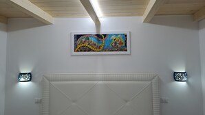 Une chambre avec un grand lit, des poutres apparentes en bois au plafond, un tableau coloré et deux lampes fixées au mur.