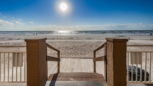 Un escalier en bois mène à une plage, sous un ciel dégagé et un soleil éclatant.