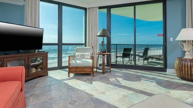 A spacious living room with a large TV, comfortable seating, and a balcony with a view of the ocean.