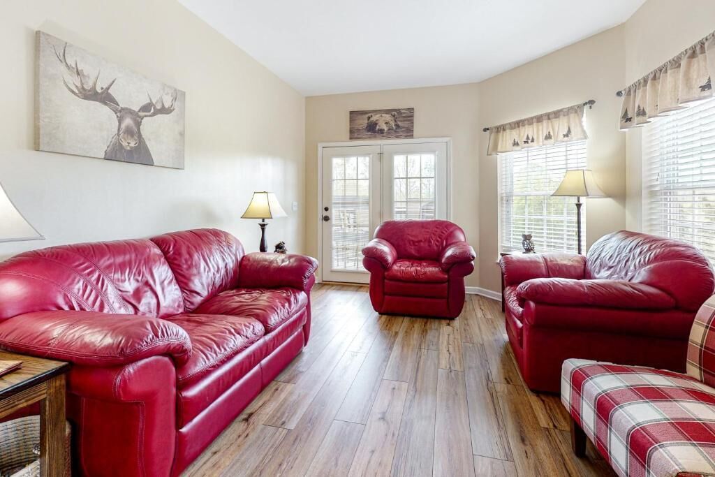 A living room with a red leather sofa, a red armchair, and a plaid ottoman. There are two lamps on the side tables, a door with a bear painting, and a deer painting on the wall.