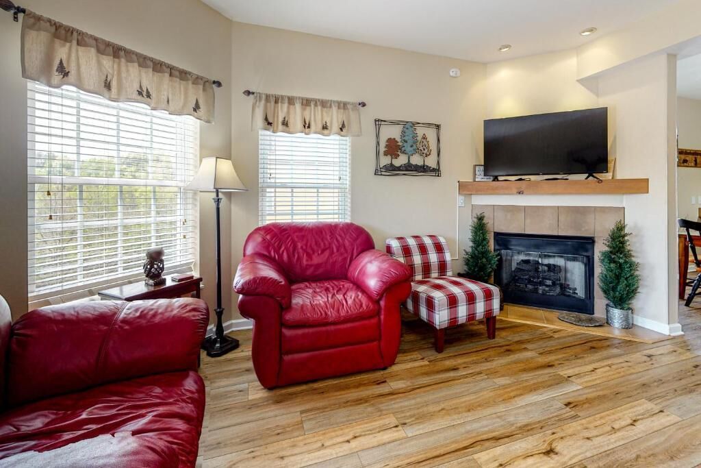 A living room with a red leather sofa, a red armchair, a plaid armchair, a fireplace, and a television on a wooden shelf.