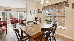 A dining area with a wooden table and chairs, a clock on the wall, and a window with blinds. - Mountain View Riverfront Condo W/ Balcony, Fishing, Fireplace, Fitness W/ Sauna (Sevierville)