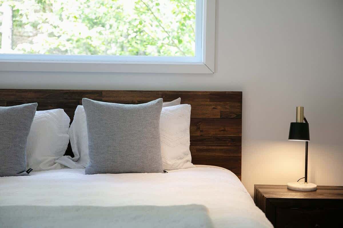 Ein ordentlich bezogenes Bett mit weißen und grauen Kissen, einem Holz-Kopfbrett und einem Nachttisch mit einer Lampe.