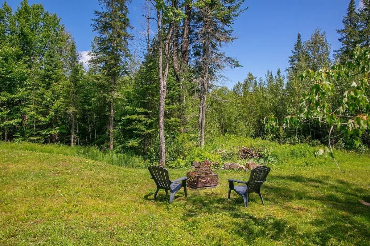 Een gazon met een buitenruimte, twee Adirondack-stoelen en een vuurkuil, omringd door bomen.