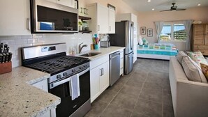 A modern kitchen with white cabinets, a stove, and a dishwasher. There is a dining area with a table and chairs, and a view of the outdoors through a window.
