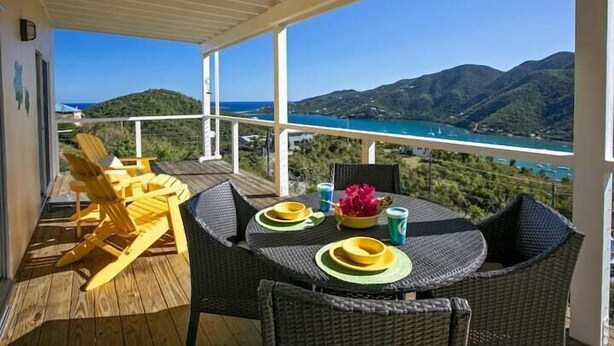 A balcony with wicker furniture, a table set with plates and cups, and a scenic view of mountains and a body of water.