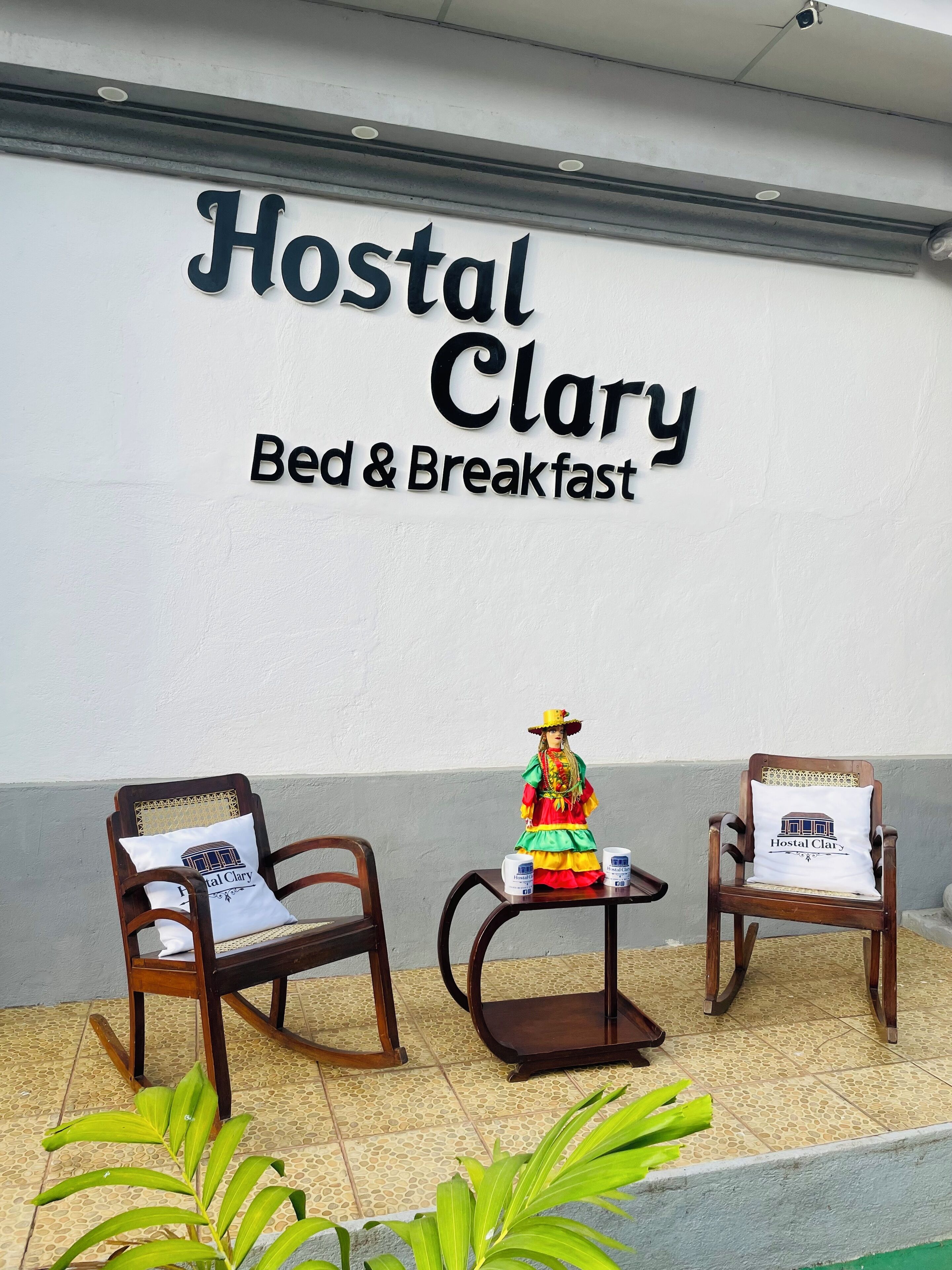 Une enseigne « Hostel Clary Bed & Breakfast », deux fauteuils à bascule en bois et une petite table sur laquelle est posée une figurine décorative.