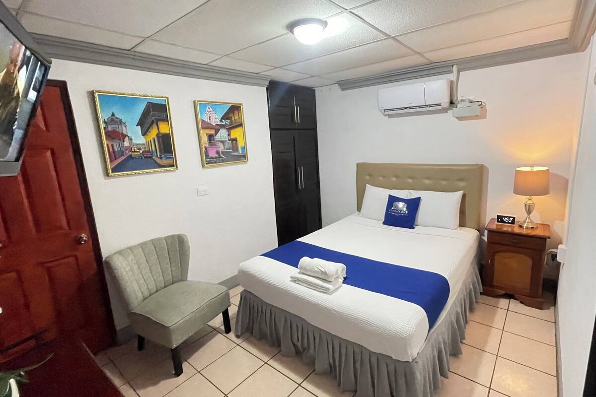 A hotel room with a bed, a chair, a desk with a lamp, and two framed pictures on the wall.