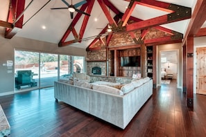 A spacious living room with a large sectional sofa, a stone fireplace, and exposed wooden beams. - Luxury 4BR Oasis | Pool & Hot Tub Near Zion (Kanab)