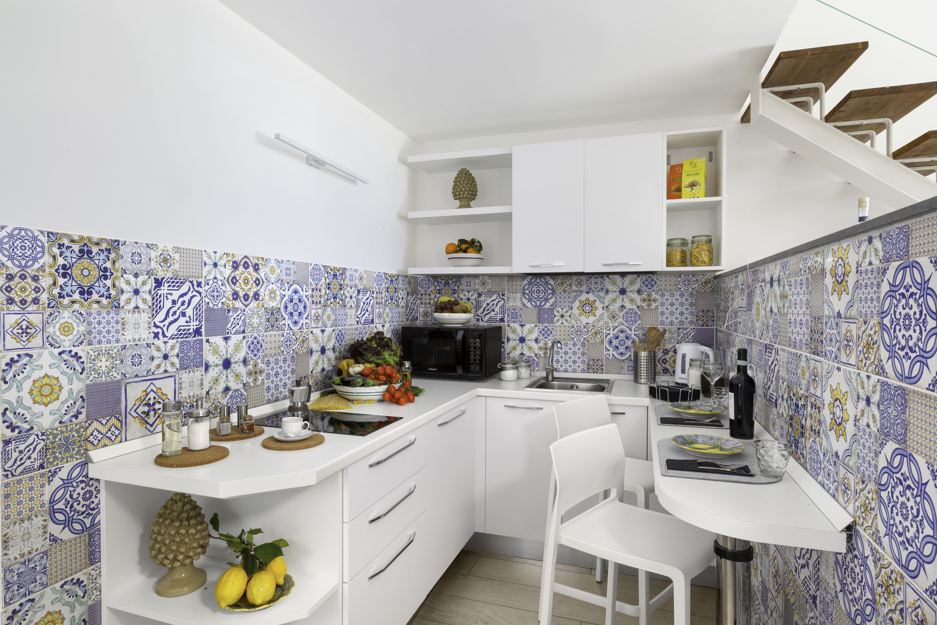 Een compacte keuken met witte kasten, een magnetron en een spoelbak. Er zijn decoratieve tegels op de wanden en een kleine fruitschaal op het aanrecht.