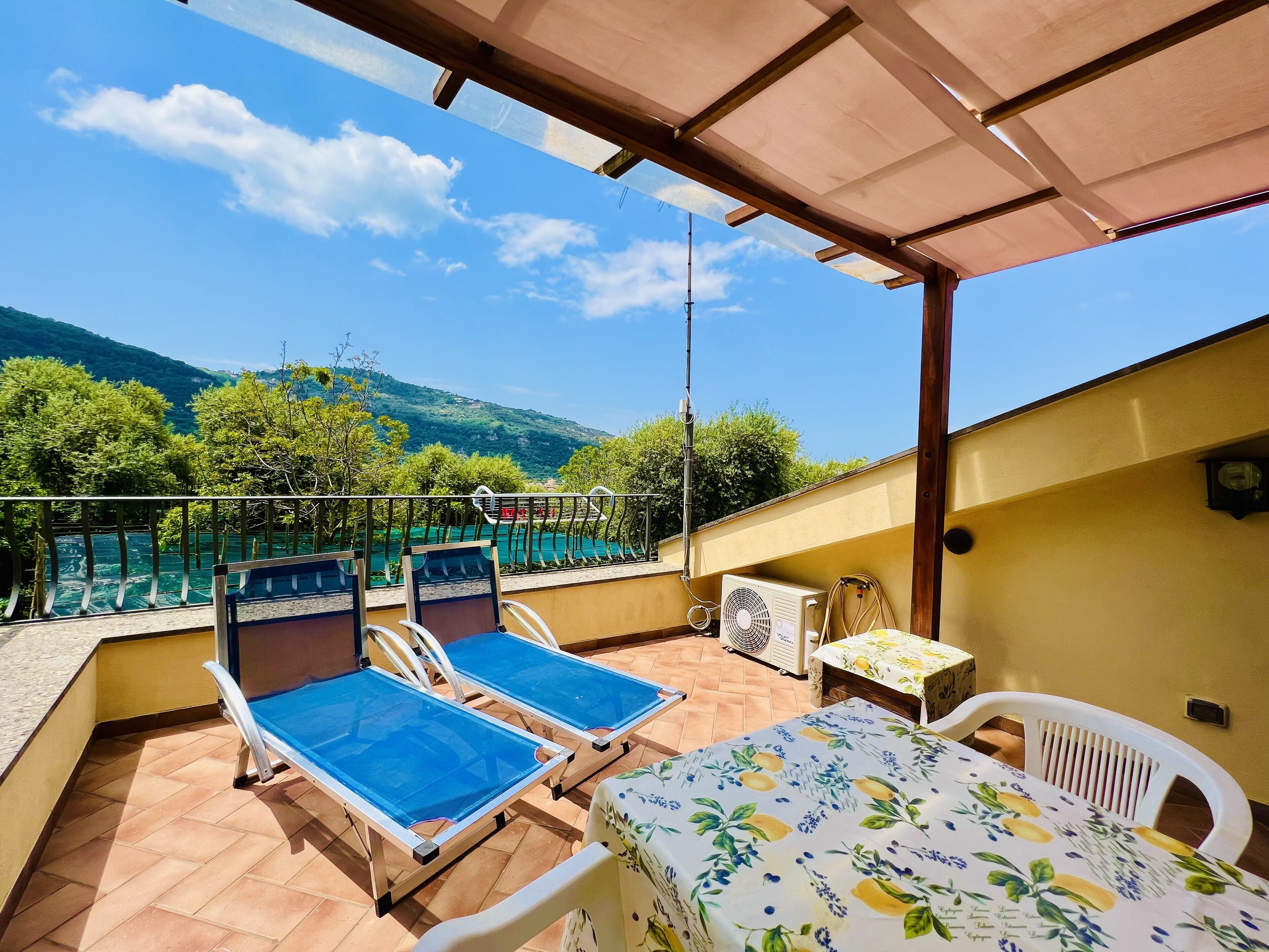 A balcony with two sun loungers, a table with a floral tablecloth, and a view of a pool and mountains.