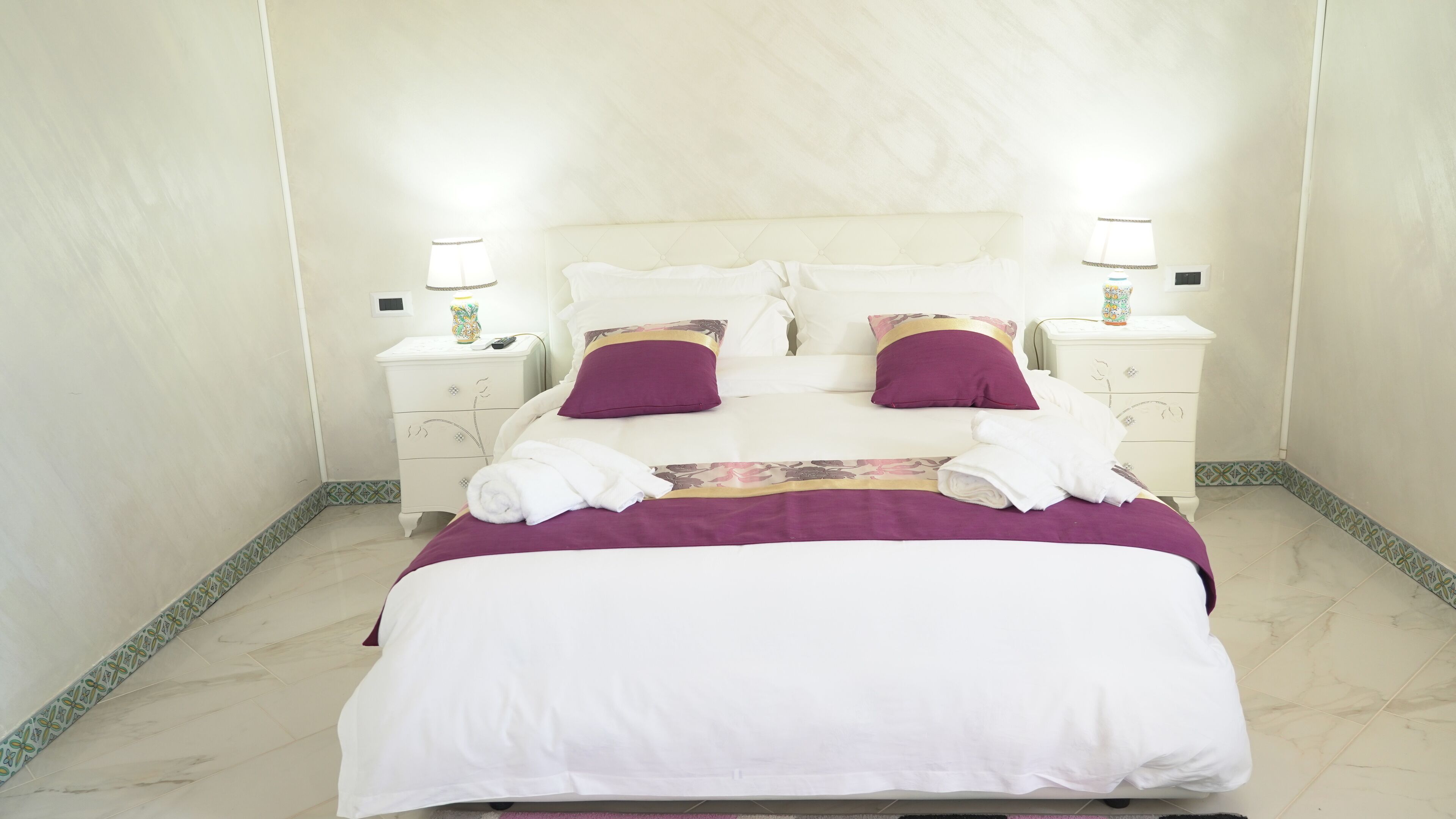 A neatly made bed with white and purple bedding, two bedside tables with lamps, and a patterned floor runner.