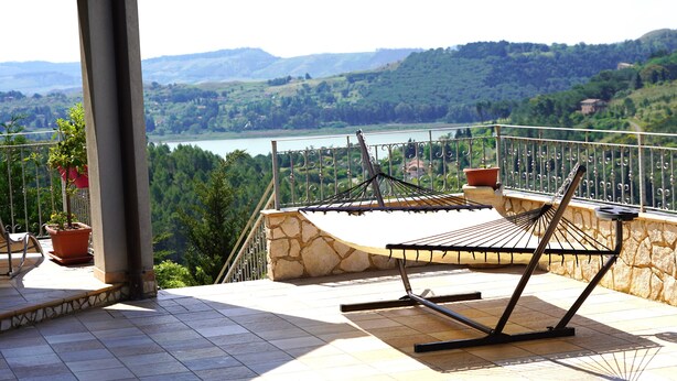 A balcony with a hammock, potted plants, and a scenic view of a lake and hills. - WE ARE LOCATED IN A VERY EXCLUSIVE AREA, WHO WOULD  KNOW?
 (Sicilia)