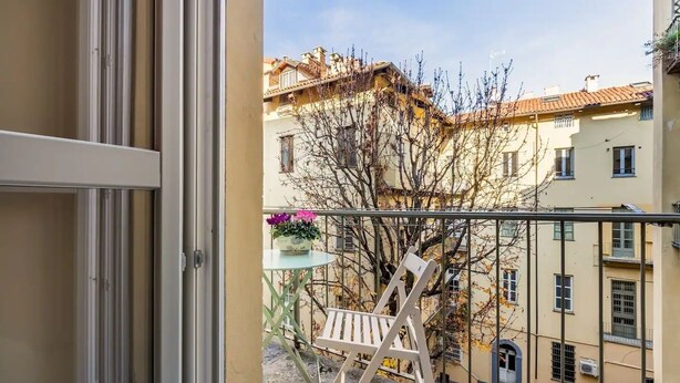 A balcony with a chair, a small table with flowers, and a view of neighboring buildings. - Santa chiara suite 2, studio in historic center  (Torino)