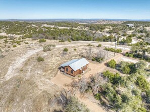 A single-story wooden cabin with a metal roof, surrounded by dry grass and sparse trees, in a remote, open landscape.