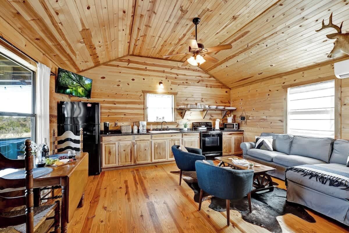 A cozy cabin interior with a wooden ceiling, a dining area, a kitchen, and a living room with a sofa and chairs.