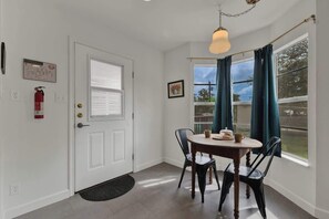 Ein Zimmer mit weißer Tür, einem Feuerlöscher, einem Fenster mit Vorhängen, einem Tisch mit Stühlen und einer Hängeleuchte.