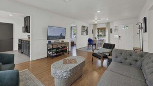 Amplia sala de estar con un sofá gris, un reposapiés estampado, un televisor de pantalla plana y un comedor con mesa y sillas.