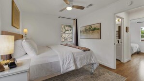 Un dormitorio con una cama, ventilador de techo, lámpara de noche, alfombra y un cuadro en la pared.
