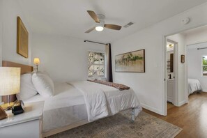 A bedroom with a bed, a ceiling fan, a bedside lamp, a window, and a door. - Stylish Abode | Minutes to Main St! (Fredericksburg)