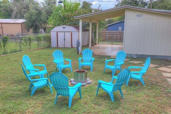 Ein Garten mit überdachter Terrasse, einem Feuerkorb und mehreren blauen Adirondack-Stühlen, die darum herum angeordnet sind.
