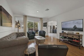 A modern living room with a grey sofa, a black leather chair, and a coffee table. There is a TV on the right wall and a painting above it. - Stylish Abode | Minutes to Main St! (Fredericksburg)