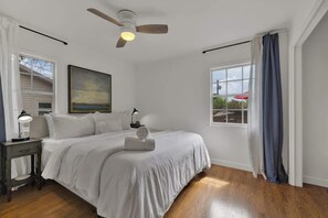 Ein Schlafzimmer mit einem Bett, einem Deckenventilator, einem Nachttisch mit Lampe, einem Bild an der Wand und einem Fenster mit Vorhängen.