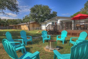 A backyard with turquoise Adirondack chairs arranged around a fire pit, a red umbrella, and a house with a deck. - Stylish Abode | Minutes to Main St! (Fredericksburg)