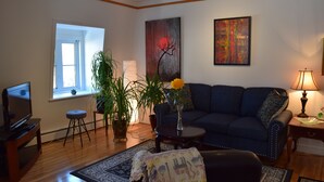 A living room with a dark blue sofa, a TV on a wooden stand, a potted plant, and a painting on the wall. - Quebec Château Richer Côte de Beaupré Canada rent a 4 ½ furnished plus more.... (Chateau-Richer)
