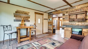 A rustic interior with exposed wooden beams, a wooden table with a chair, a checkered rug, and a kitchen area with wooden cabinets and a refrigerator.