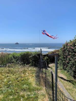 A coastal view with a kite flying over the ocean, a fence, and a bush with pink flowers. - Sunset Shore Retreats Ocean Front "Cozy Seal Slumber" (Smith River)