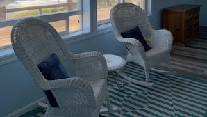 Een kamer met twee rieten stoelen, een houten kast en uitzicht op de oceaan door grote ramen.