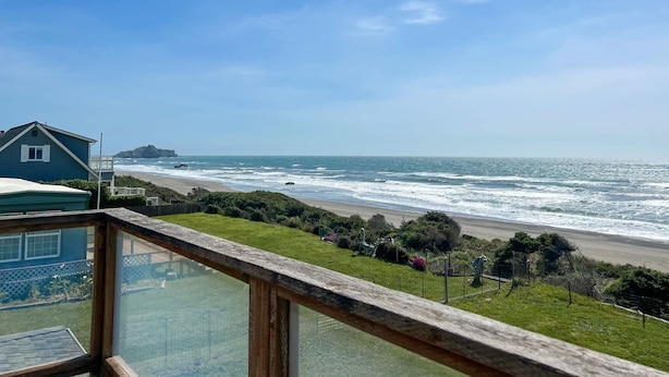 Uitzicht op de kust vanaf een balkon, met heldere lucht, golven en een verafgelegen rotsformatie.