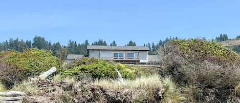 A coastal house with a sandy foreground, dense vegetation, and a clear blue sky.