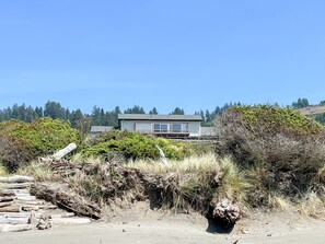 A coastal house with a sandy foreground, dense vegetation, and a clear blue sky.