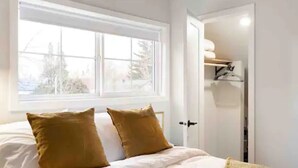 Een slaapkamer met een groot raam, een bed met witte beddengoed en twee mosterdkleurige kussens, en een deur naar een inloopkast met planken.