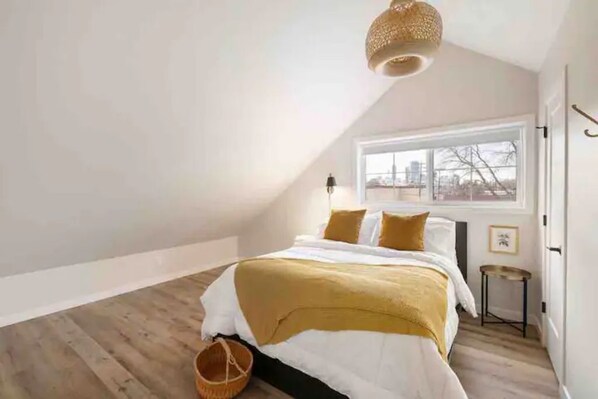 Un dormitorio con un ventanal grande, una cama con una manta amarilla, una lámpara colgante y suelo de madera.