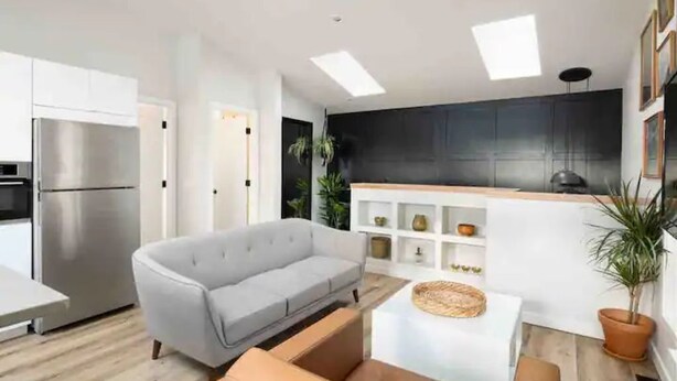 Een moderne keuken met witte kasten, een roestvrijstalen koelkast en een zwart aanrecht. Er staat een grijze bank en een kleine tafel met een rieten mandje.
