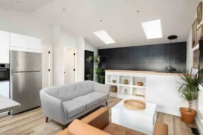 A modern kitchen with white cabinets, a stainless steel refrigerator, and a black countertop. There is a gray sofa and a small table with a woven basket.