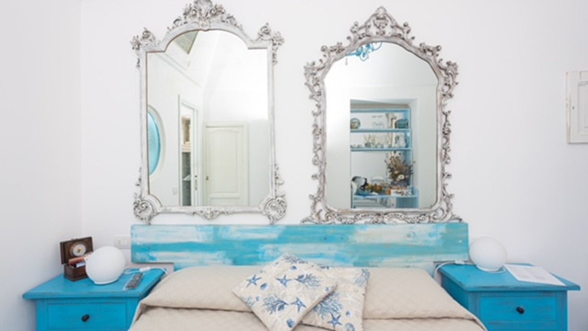 A bedroom with two ornate mirrors, a blue dresser, a bed with a floral pillow, and a shelf with decorative items.