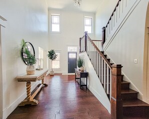 Un hall d’entrée spacieux avec un escalier en bois, une console agrémentée d’un miroir et de plantes, et une porte dotée d’une vitre en verre dépoli.