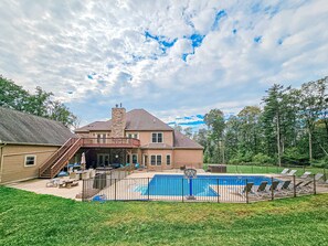Une maison à deux étages avec une cheminée en pierre, un balcon et une piscine entourée d’une clôture.