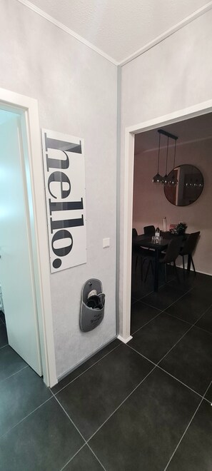Käytävässä on kyltti, jossa lukee ”hei”, kenkäteline sekä ruokailutila, jossa on pyöreä peili ja riippuvalaisimet.
